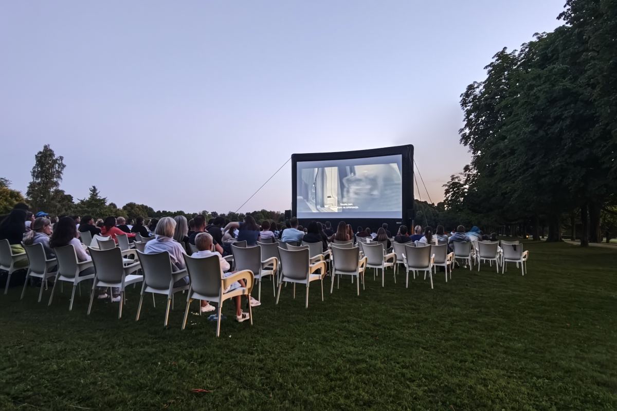 Filmski večer v mraku Obiskovalci sedijo na stolih pred velikim filmskim platnom, na katerem se predvaja film, nad parkom se spušča mrak.