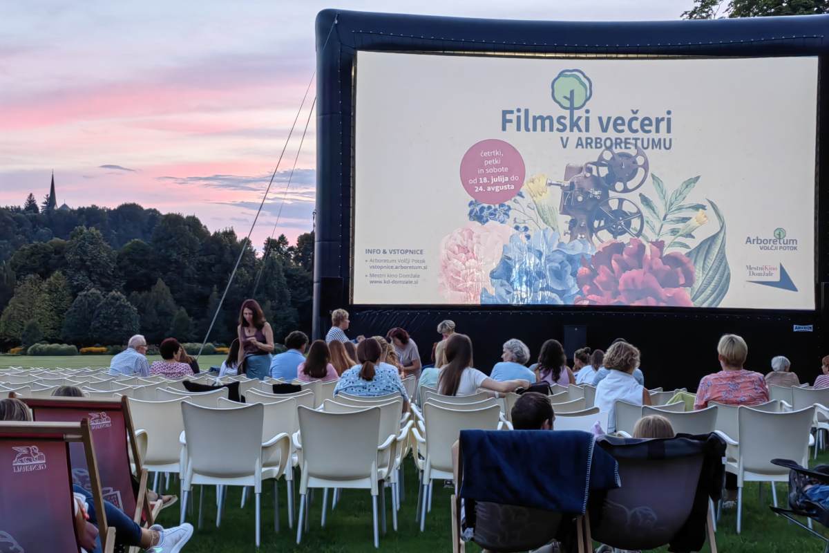 Filmski večeri v Arboretumu Obiskovalci sedijo pred velikim platnom z napisom Filmski večeri v Arboretumu, v ozadju je rožnato večerno nebo.