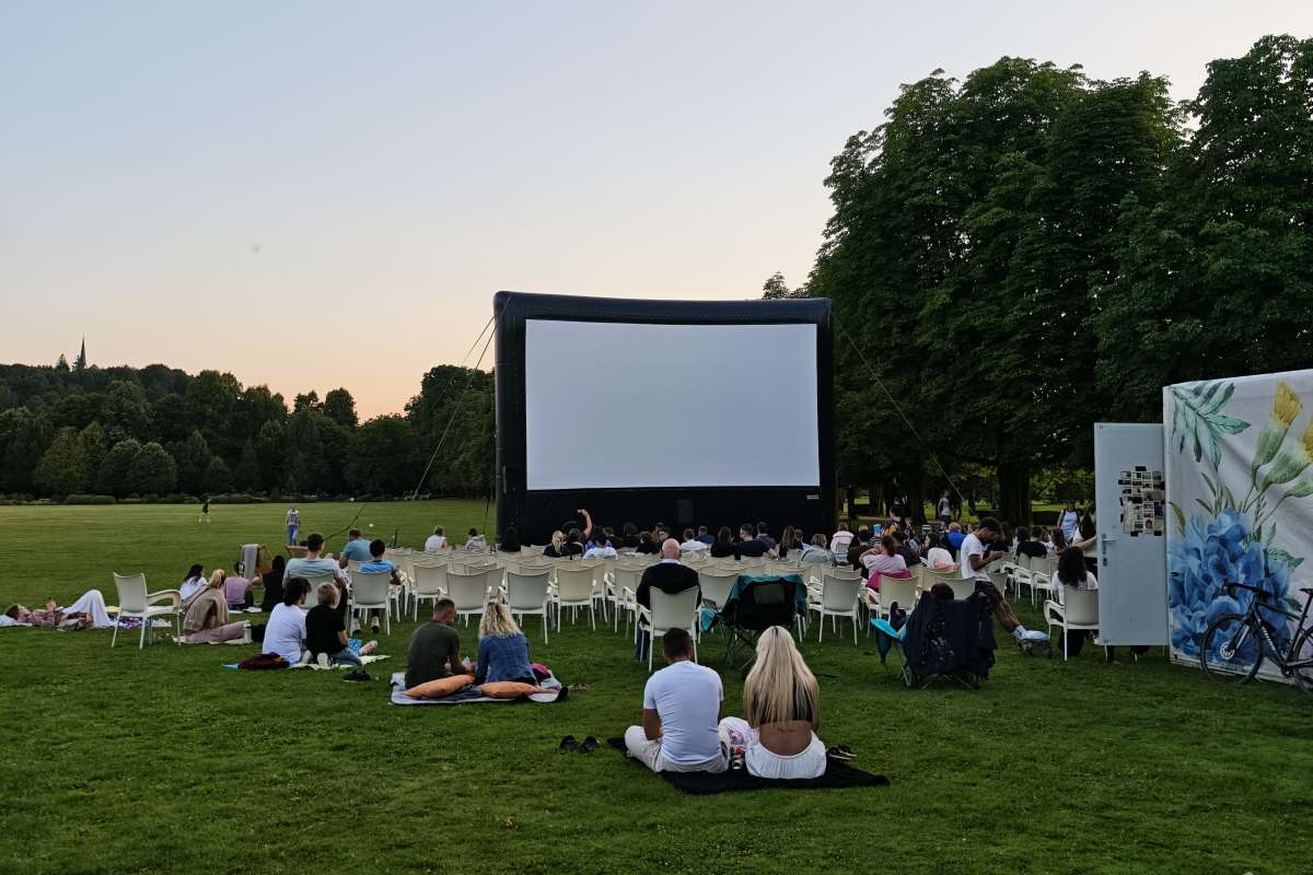 Filmski večer v Arboretumu Obiskovalci sedijo na travniku in na stolih pred velikim filmskim platnom v Arboretumu ob večernem nebu.