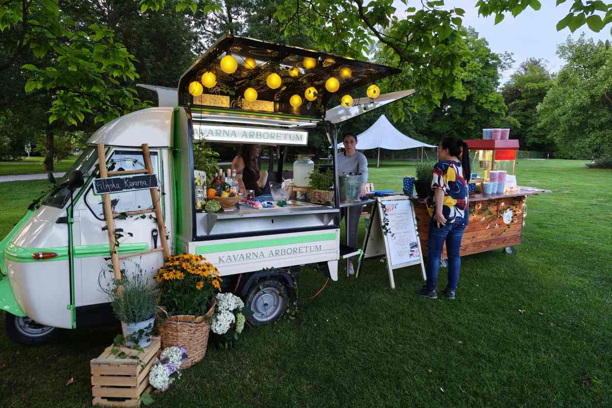 Kavarna pod drevesi Mobilna kavarna Arboretum stoji na travniku pod drevesi, ob njej obiskovalci izbirajo pijačo in prigrizke pred filmskim večerom.
