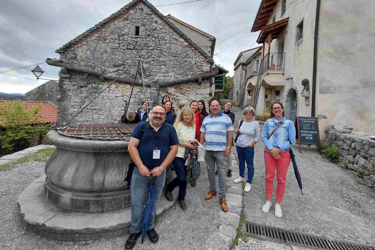 Obiskovalci ob kraškem vodnjaku v Štanjelu Skupina obiskovalcev stoji ob kamnitem vodnjaku v starem jedru Štanjela. V ozadju so kamnite hiše, ozka kraška ulica in oblačno nebo.