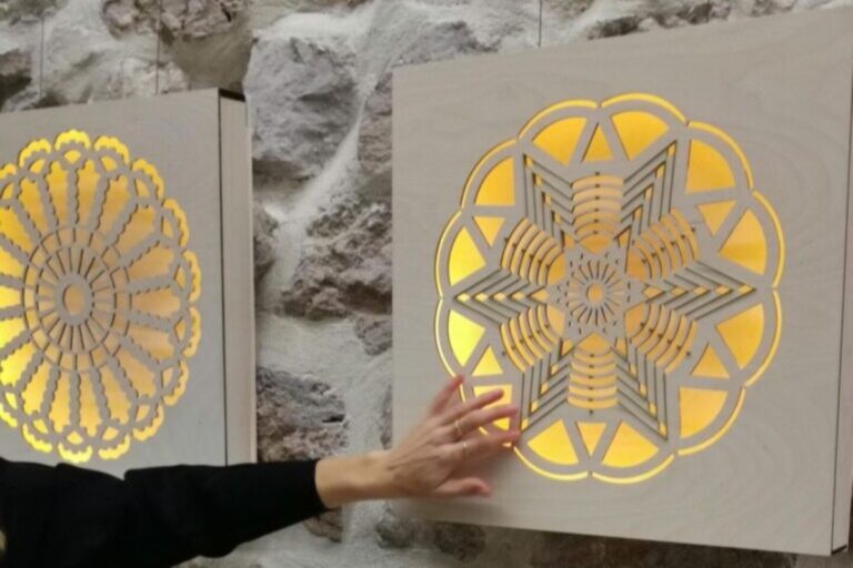 Oseba z roko tipa osvetljeno taktilno skulpturo z geometrijskim vzorcem, nameščeno na steni. V ozadju je kamnita stena, levo pa je vidna še druga podobna osvetljena skulptura.