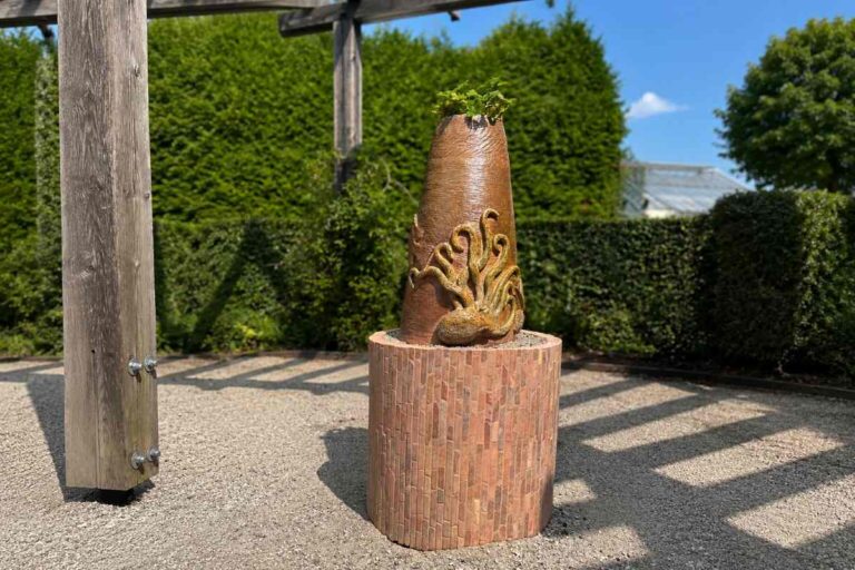 Skulptura z reliefom hobotnice stoji na okroglem podstavku iz mozaika. Na vrhu skulpture raste zeleno rastlinje, v ozadju pa so živa meja, lesena pergola in jasno modro nebo v sončnem vremenu.