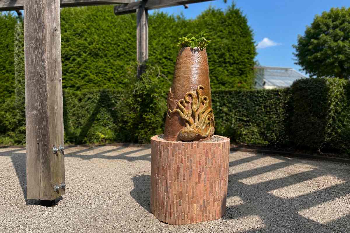 Skulptura z reliefom hobotnice stoji na okroglem podstavku iz mozaika. Na vrhu skulpture raste zeleno rastlinje, v ozadju pa so živa meja, lesena pergola in jasno modro nebo v sončnem vremenu.
