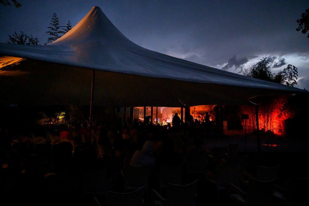 Večerni ambient Cvetličnega orkestra. Foto: Katja Goljat Pogled na Indonezijski paviljon v večernem mraku, kjer obiskovalci spremljajo Cvetlični orkester ob svetlobni osvetlitvi in temnih nevihtnih oblakih.