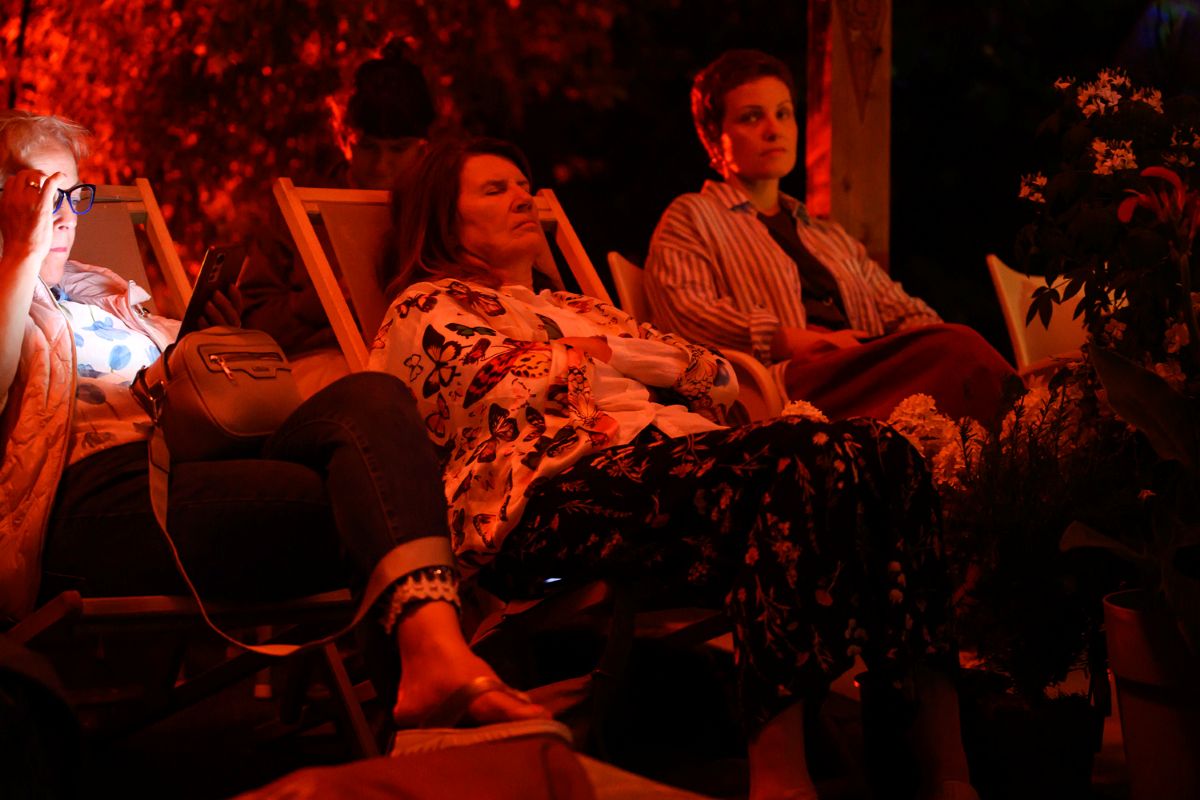 Obiskovalci prisluhnejo Cvetličnemu orkestru Foto: Katja Goljat Obiskovalci sproščeno sedijo in ležijo na ležalnikih pod Indonezijskim paviljonom ter poslušajo zvočni performans v rdeče osvetljenem večernem ambientu.
