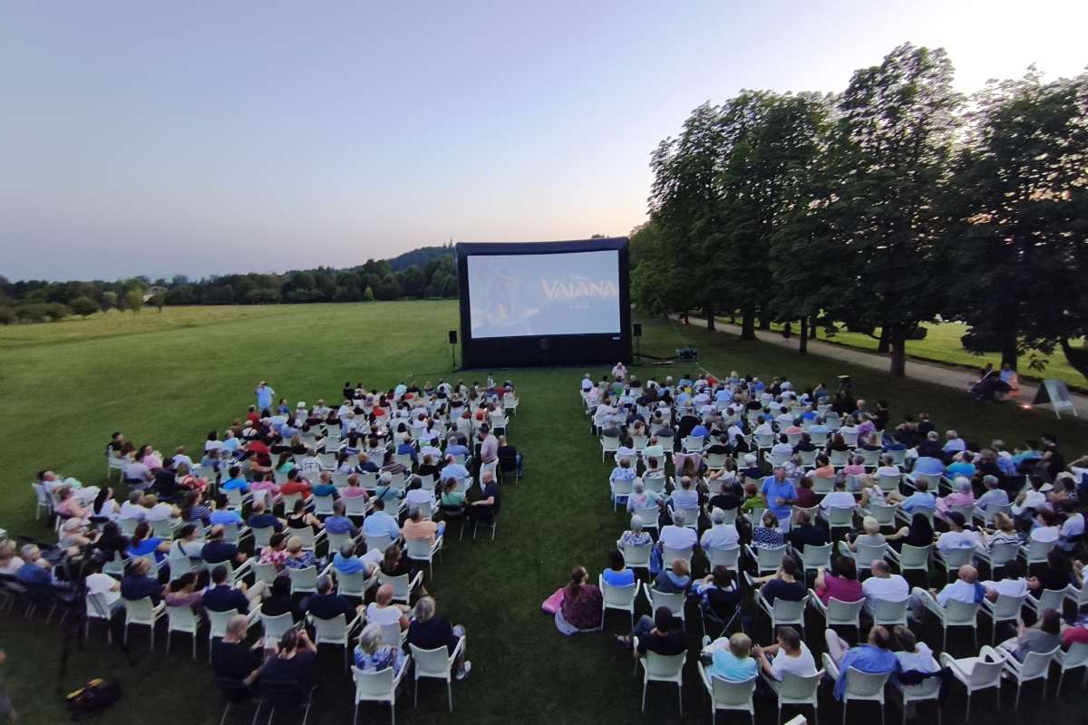 Filmski večeri pod zvezdami v Arboretumu Širok pogled na filmsko prizorišče na travniku v Arboretumu. Pred velikim platnom sedi številno občinstvo na belih stolih, na zaslonu pa je prikazan naslov animiranega filma Vaiana. V ozadju so drevoredi in večerno nebo.