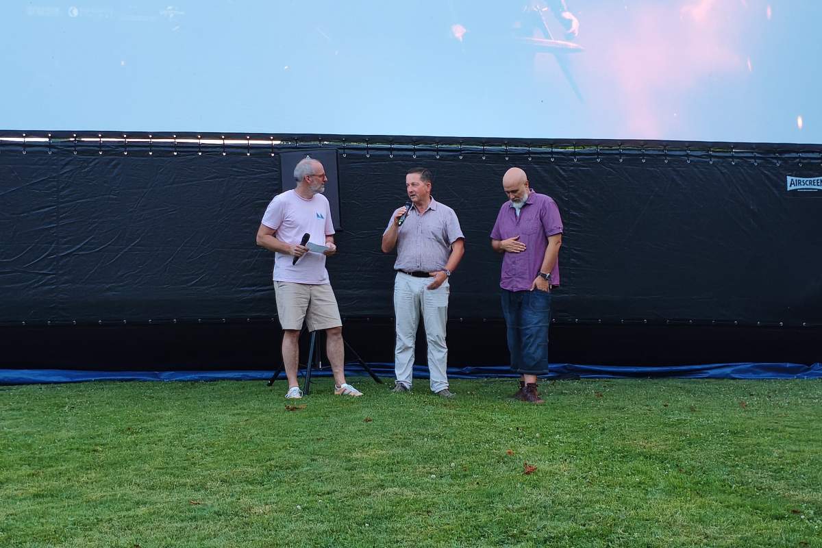 Uvodni nagovor pred premierno projekcijo filma Fotograf Trije moški stojijo pred velikim napihljivim filmskim platnom na travniku. Eden govori v mikrofon, druga dva stojita ob njem in poslušata. Dogodek poteka na prostem tik pred začetkom večerne filmske projekcije.