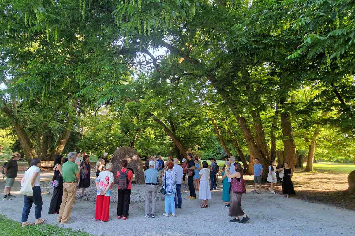 Vodeni ogled taktilne razstave skulptur v Arboretumu Volčji Potok Obiskovalci med vodenim ogledom stojijo okoli taktilne skulpture pod krošnjami dreves v Arboretumu Volčji Potok.