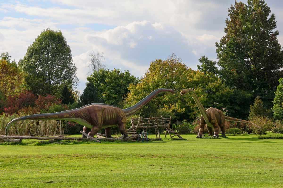Dinozavri med jesenskimi barvami. Foto: Klara Kopač. Dve veliki maketi dolgovratih dinozavrov stojita na zeleni jasi, obdani z drevesi v jesenskih barvah.