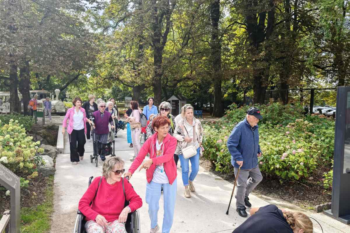 Obiskovalci Doma upokojencev Kranj na Poti za spomin Obiskovalci Doma upokojencev Kranj se s spremljevalci sprehajajo po široki poti med cvetočimi hortenzijami. Nekateri uporabljajo invalidski voziček, hoduljo ali palico.