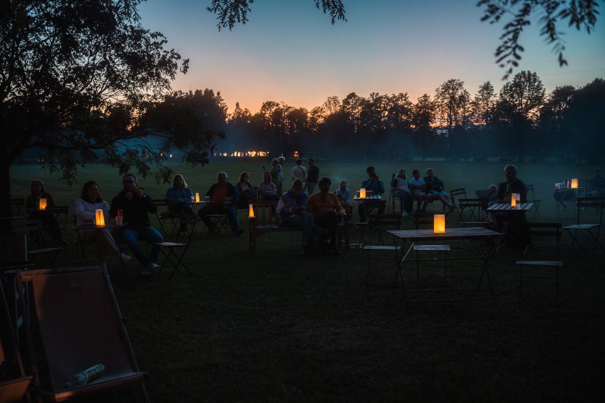 Cvetlični orkester ob sončnem zahodu, FOTO: Tadej Hrastovec. Obiskovalci ob mraku sedijo za mizami s prižganimi lučkami, za drevesi pa se nebo obarva oranžno.