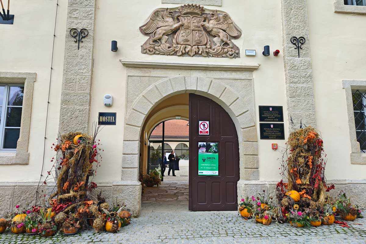 Jesenska okrasitev vhoda v dvorec Opeka Kamnit obokan vhod v dvorec Opeka z grbom nad vrati ter jesenskima aranžmajema iz buč, cvetja in vej ob straneh.