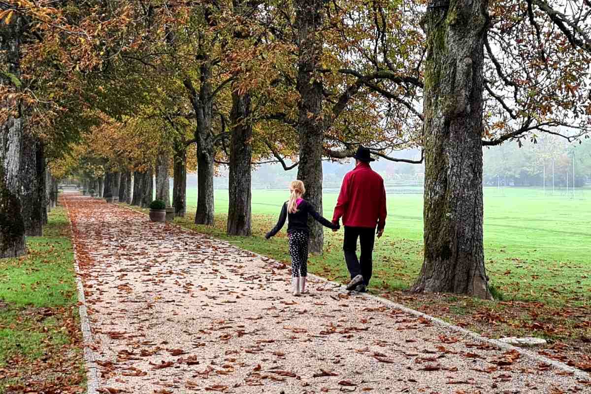 Jesenski sprehod skozi drevored v Arboretumu Otrok in odrasla oseba v rdeči jakni se držita za roke med sprehodom po drevoredu. Peščeno pot prekriva odpadlo rjavo listje.