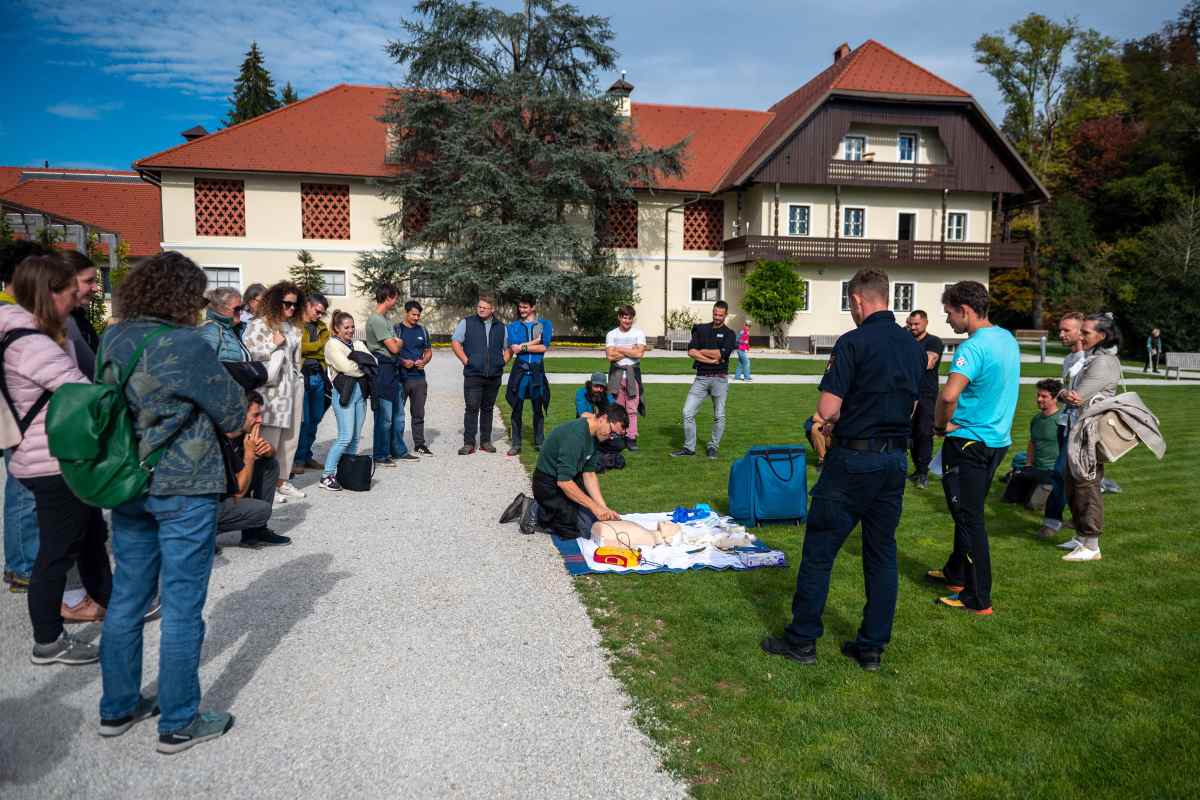 Udeleženci na zelenici opazujejo prikaz oživljanja na lutki, ob kateri so razporejeni pripomočki za prvo pomoč.