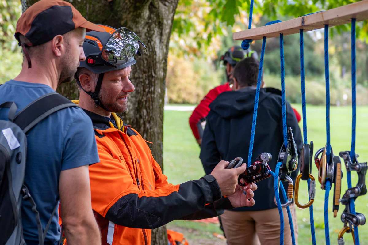 Predstavitev pripomočkov za plezanje. Foto: Nika Halaš Arborist v oranžni zaščitni obleki udeležencu kaže plezalni pripomoček. Ob njima na modrih vrveh visijo vponke in druge naprave.