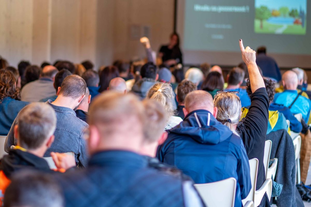 Vprašanja in razprava med strokovnimi predavanji. Foto: Nika Halaš Udeleženka v polni predavalnici dviguje roko, medtem ko drugi poslušajo predavateljico pred projekcijskim platnom.