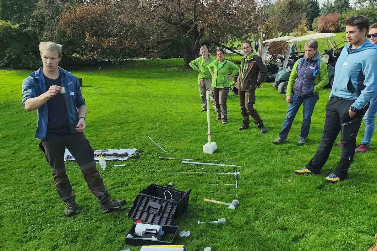 Ena od terenskih delavnic v sklopu Slovenskih arborističnih dni 2026. Foto: Nika Halaš Izvajalec na zelenici skupini kaže kovinski valj. Na travi pred njim so razporejeni pripomočki za vzorčenje tal.