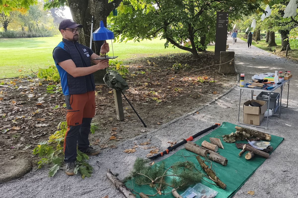 Terenska delavnica v sklopu Slovenskih arborističnih dnevov 2026, Foto: Nika Halaš Izvajalec ob drevesu drži modro lovilno past. Na podlogi pred njim so vzorci vej in lesa, na mizi pa pripomočki.