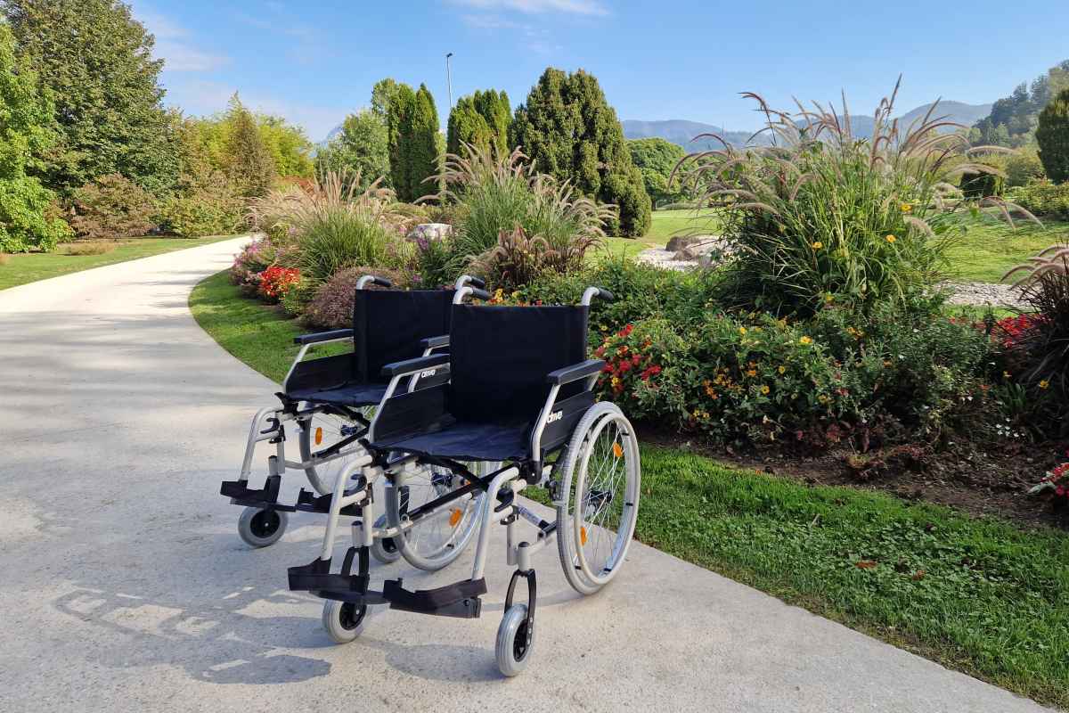 Dva invalidska vozička na utrjeni poti ob cvetličnih gredah in okrasnih travah v Arboretumu Volčji Potok.