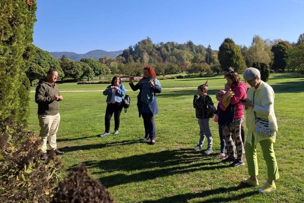 Matjaž Mastnak o vplivu podnebnih sprememb na iglavce Direktor Matjaž Mastnak stoji ob visokem iglavcu in razlaga skupini obiskovalcev, ki ga poslušajo na zelenici Arboretuma Volčji Potok.