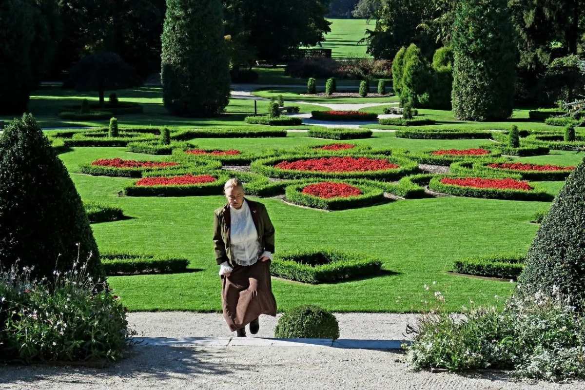 Po sledeh Helene Souvan v Arboretumu Ženska v dolgem rjavem krilu in temnem suknjiču se vzpenja po kamnitih stopnicah, za njo pa se razprostira francoski vrt z rdečimi cvetličnimi gredicami.
