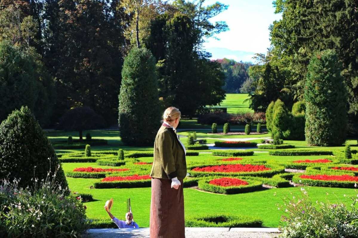 Helena Souvan ob pogledu na francoski vrt Ženska v zgodovinskih oblačilih stoji na robu francoskega vrta in opazuje geometrijske cvetlične gredice, medtem ko moški v ozadju dviguje slamnik.